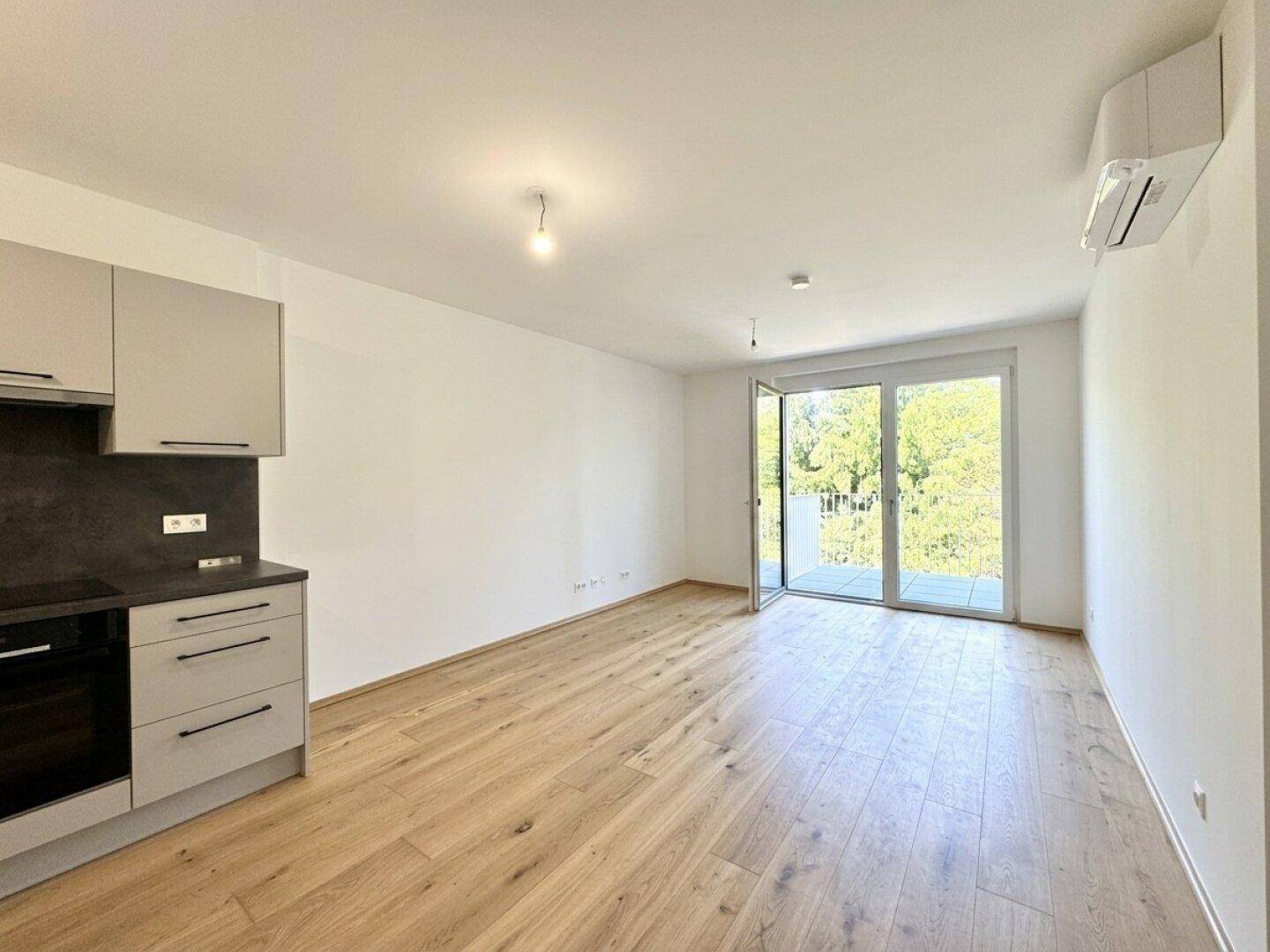 Heller offener Wohnbereich mit integrierter Küchenzeile, Holzboden und Zugang zum Balkon mit Grünblick.