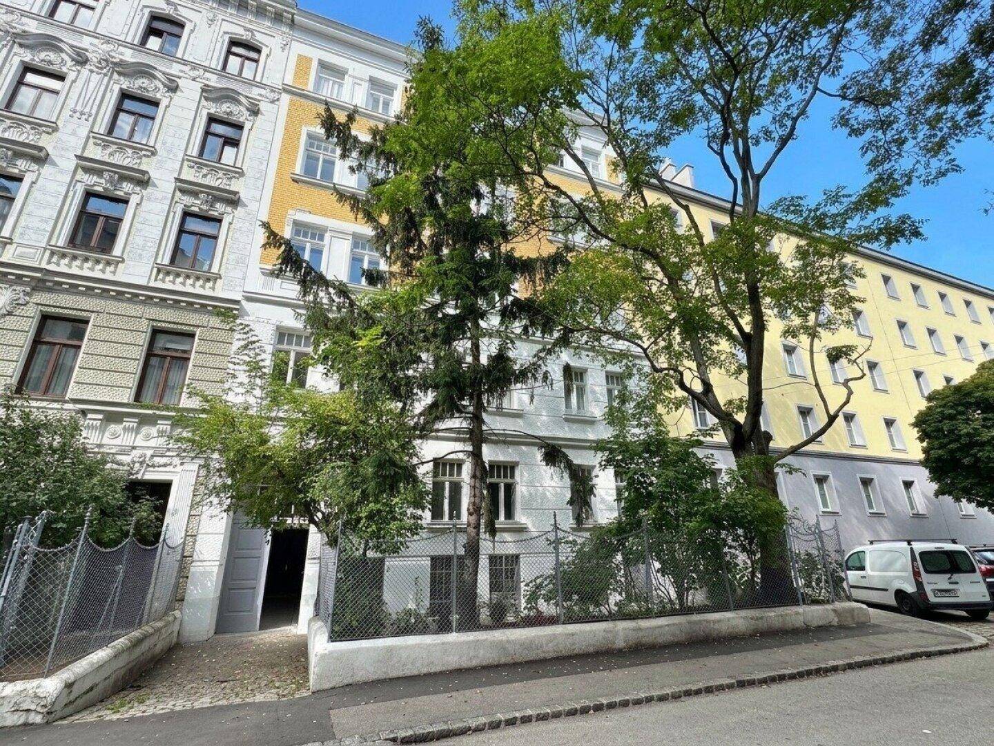 Historisches Mehrfamilienhaus mit klassischer Fassade und hohen Bäumen an der Straße.