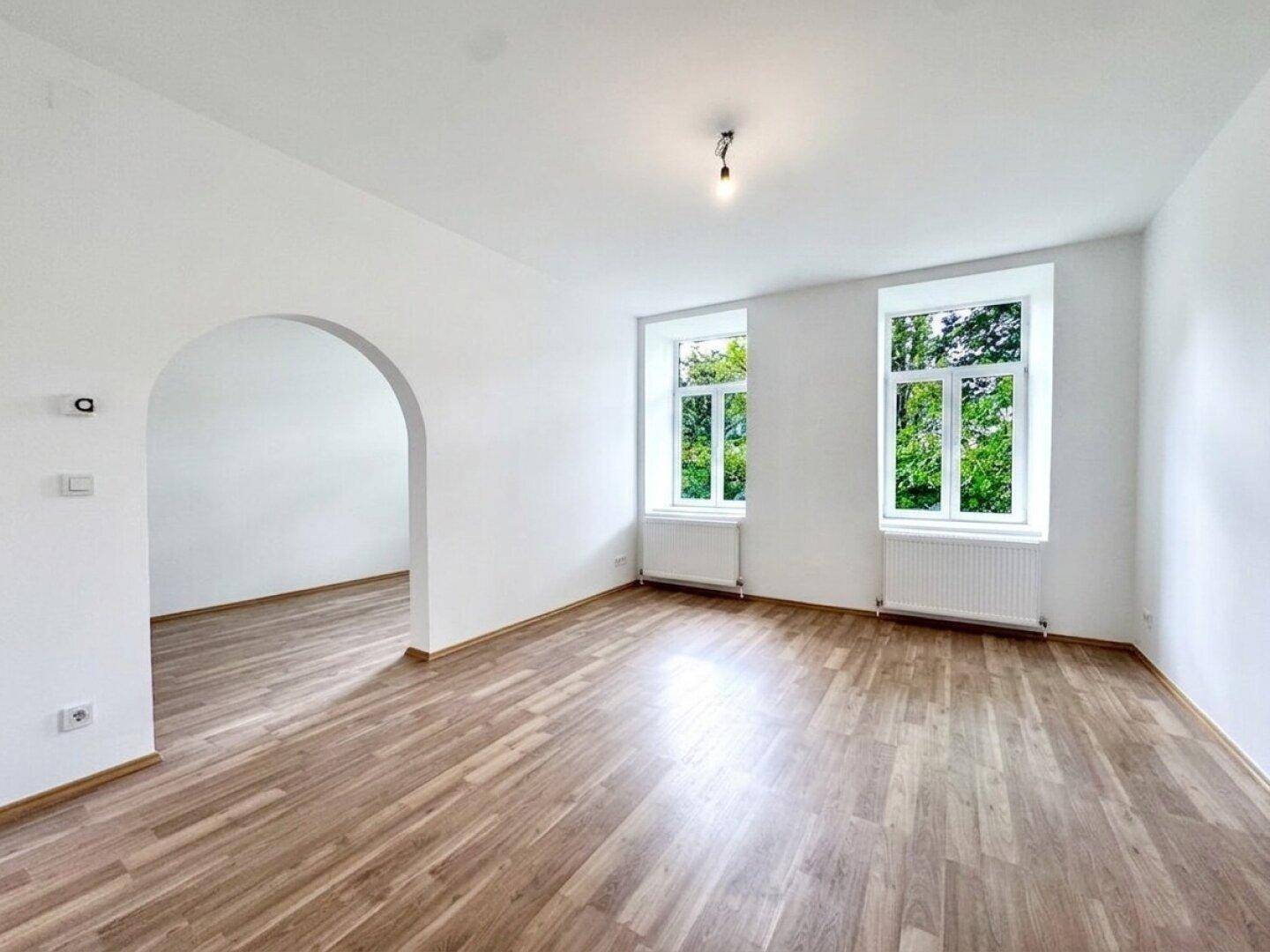 Geräumiges, unmöbliertes Wohnzimmer mit Holzboden und zwei großen Fenstern, die viel Tageslicht hereinlassen.