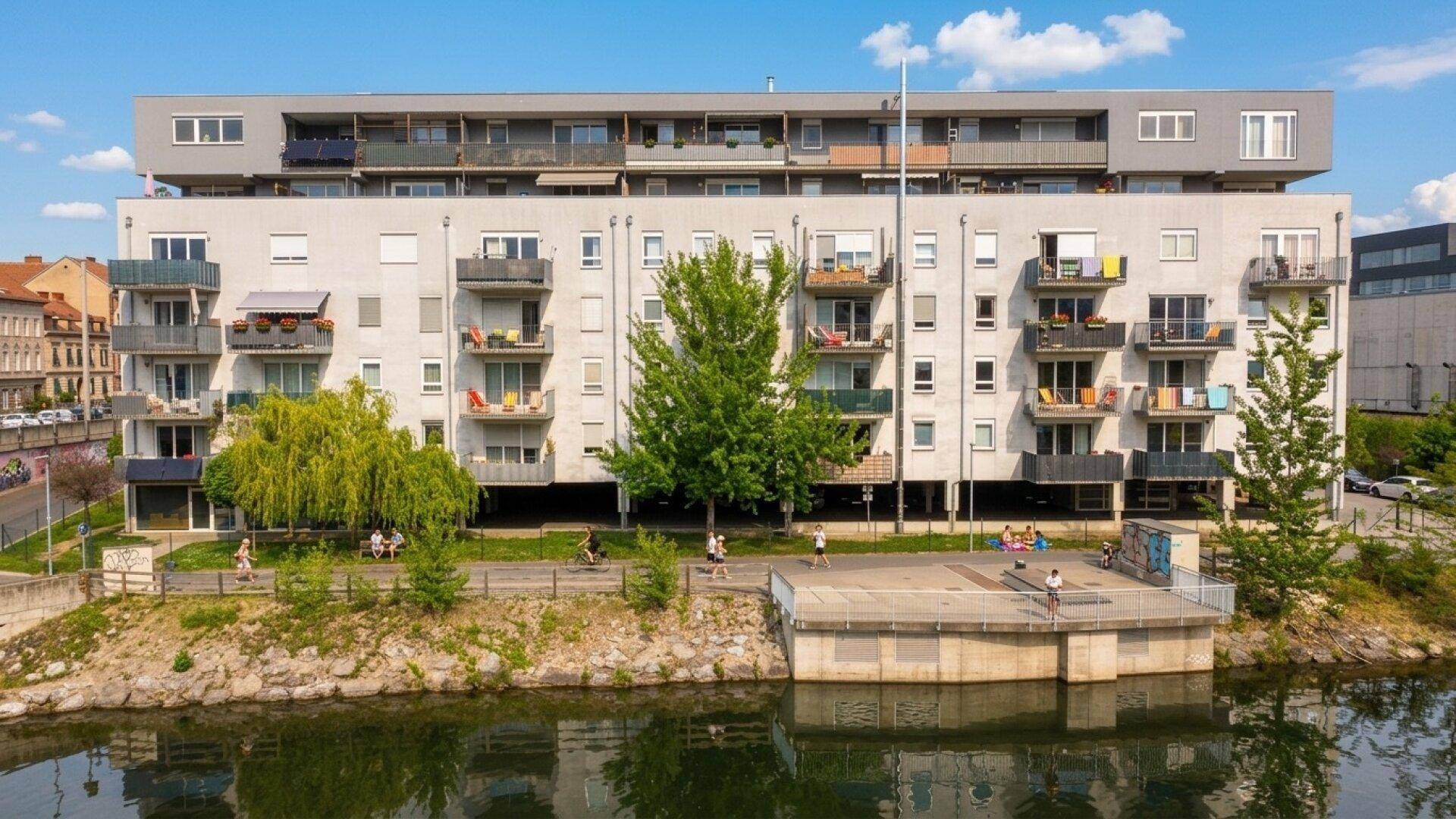 Modernes Mehrfamilienhaus am Flussufer mit zahlreichen Balkonen und Grünflächen.