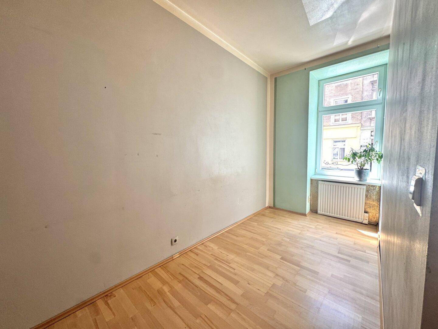 Leeres Schlafzimmer mit Parkettboden, hellem Fenster und Heizkörper unter dem Fenster.