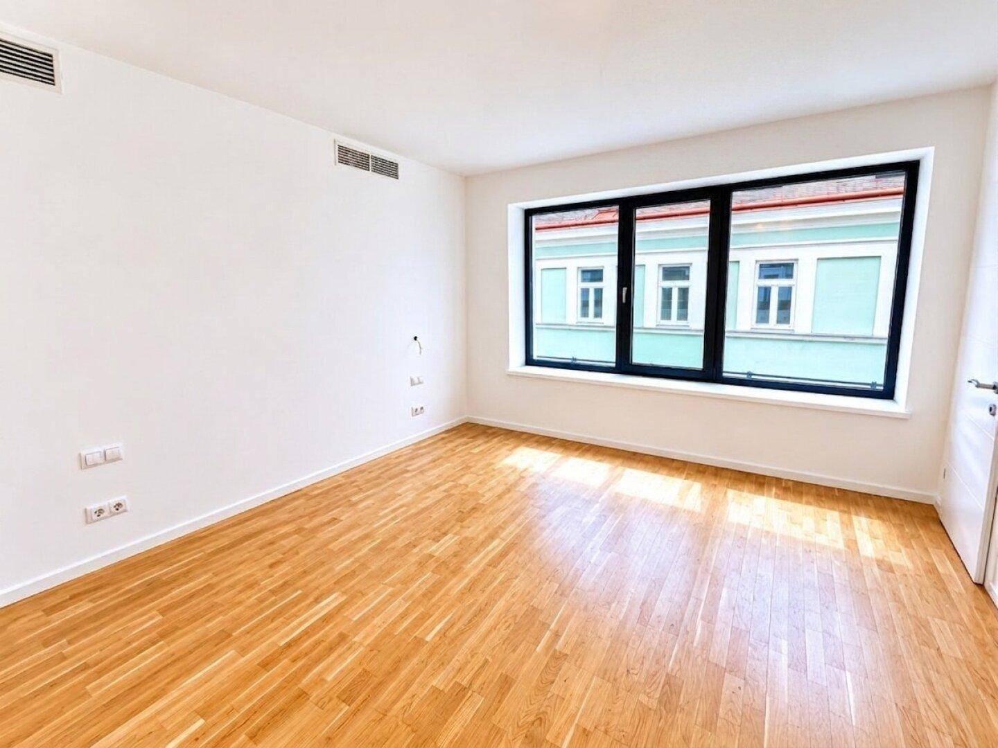 Geräumiges, helles Zimmer mit Holzboden und einem Fenster mit Blick auf die gegenüberliegende Fassade.