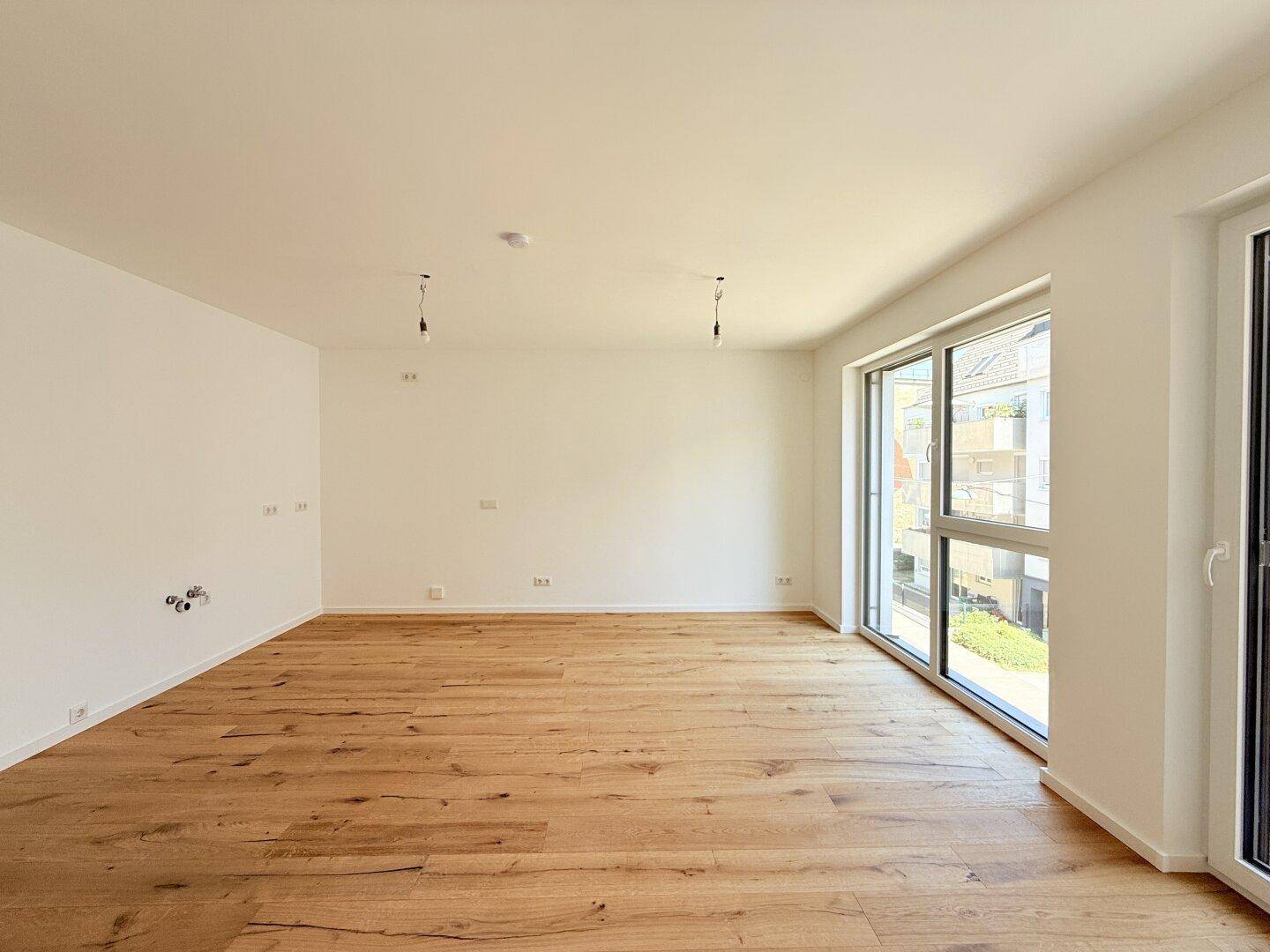 Geräumiges Wohnzimmer mit hellem Holzboden und großen Fenstern, die einen Blick nach draußen bieten.