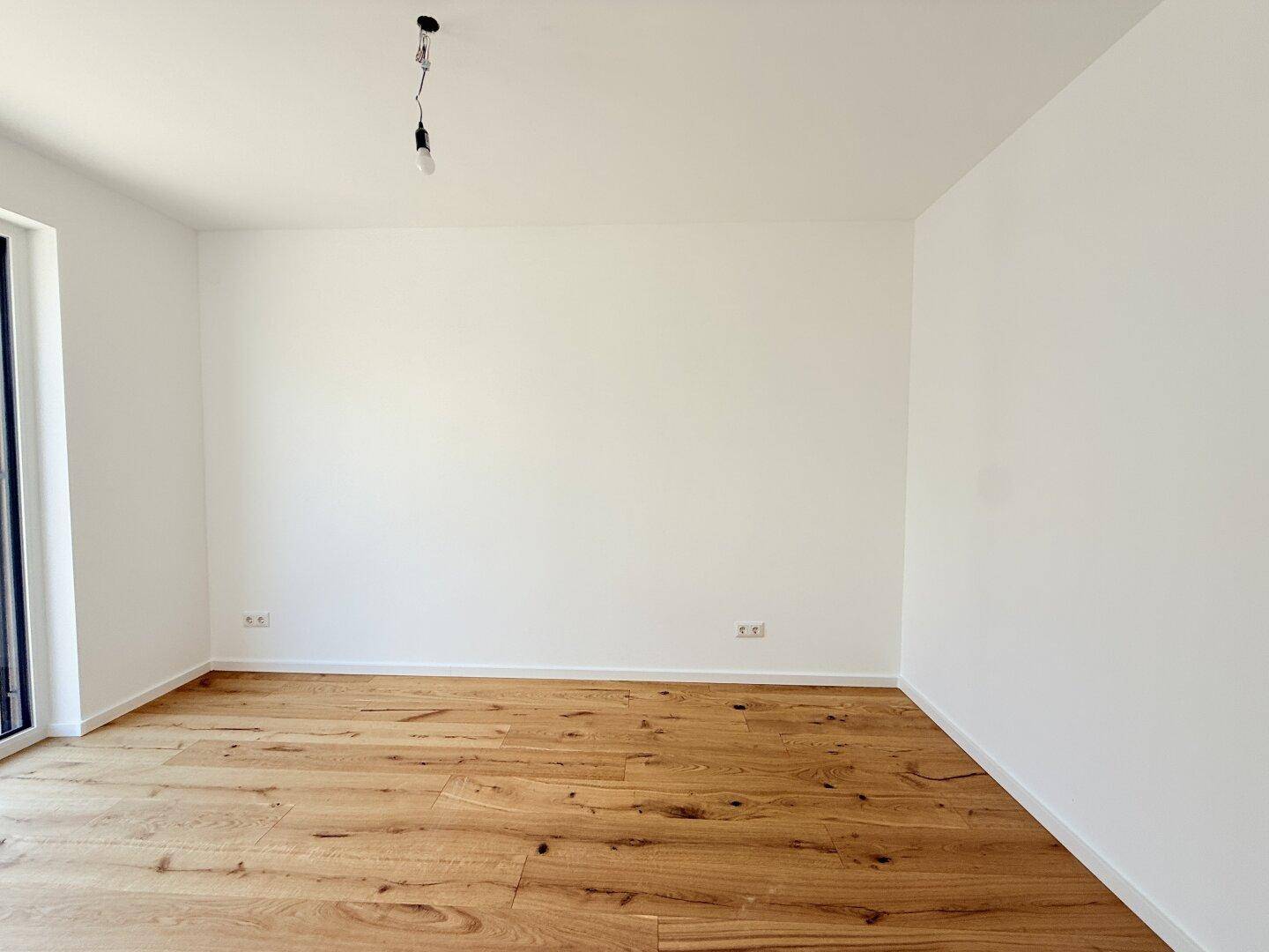 Leeres Schlafzimmer mit Holzboden und einem großen Fenster, das für viel Helligkeit sorgt.