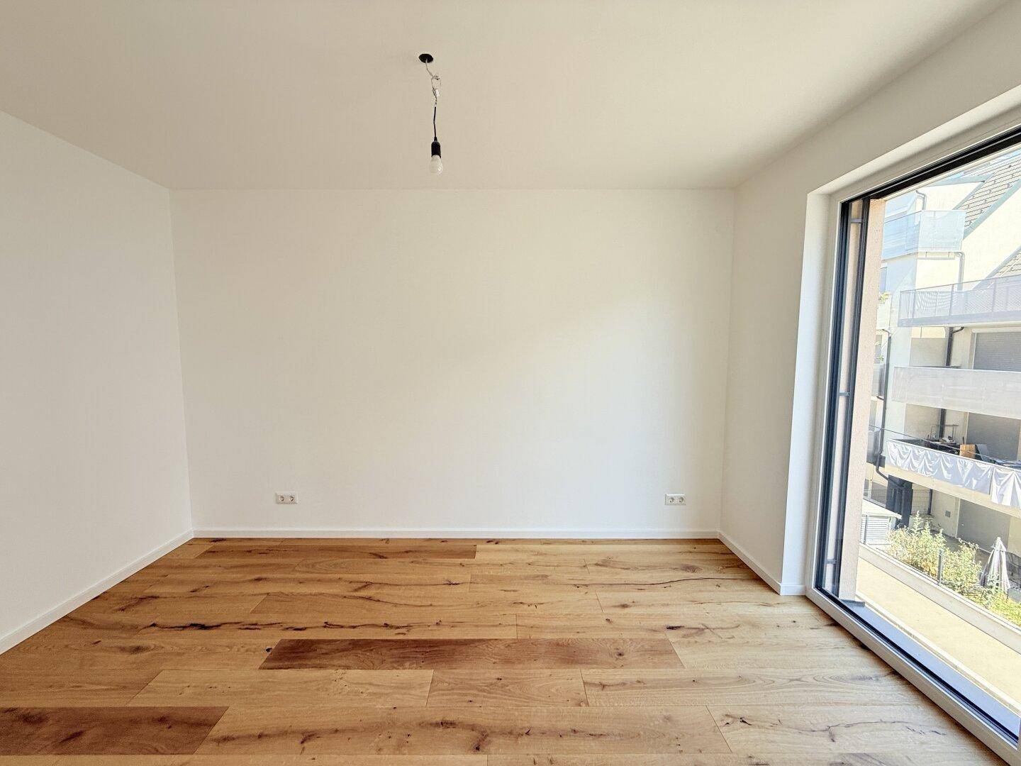 Leeres Zimmer mit Holzboden und großem Fenster, das viel natürliches Licht in den Raum bringt.