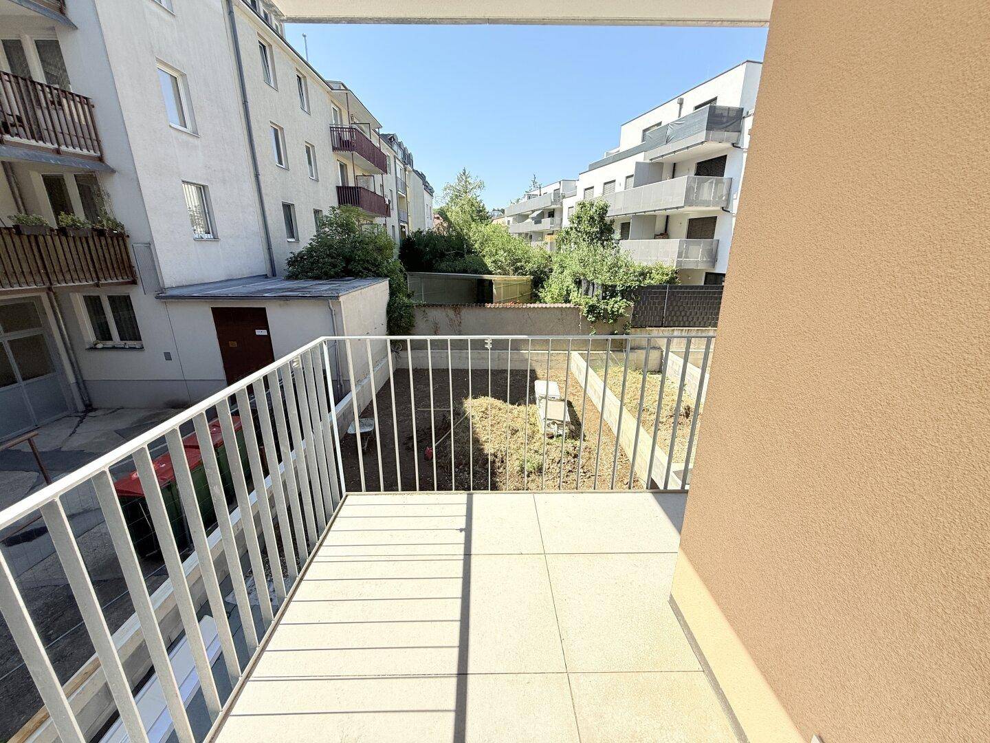 Heller Balkon mit Fliesenboden und Blick auf den Innenhof und umliegende Gebäude bei Sonnenschein.