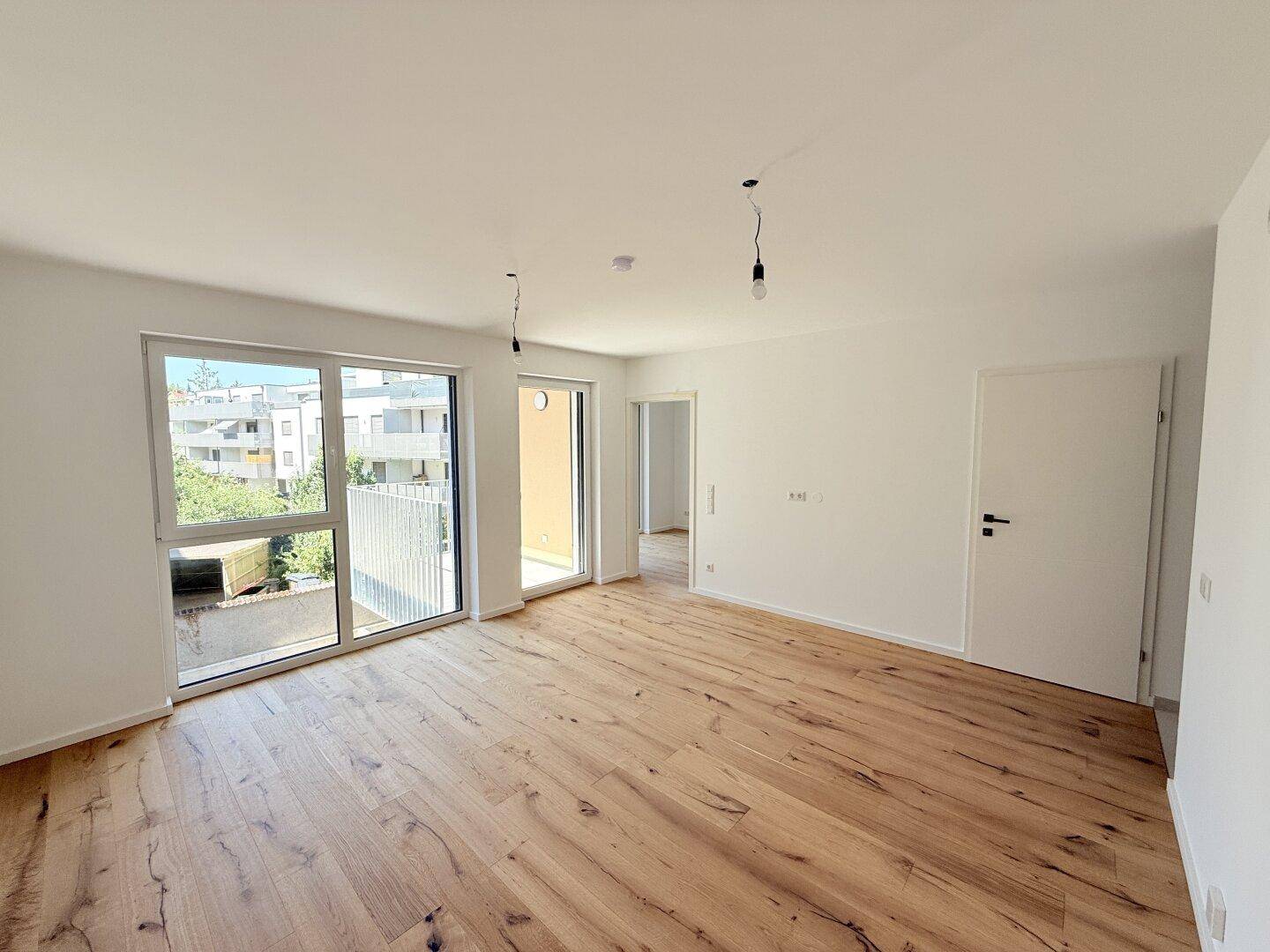 Geräumiges Zimmer mit Holzboden und großen Fenstern, die viel Tageslicht hereinlassen und einen Balkon zeigen.