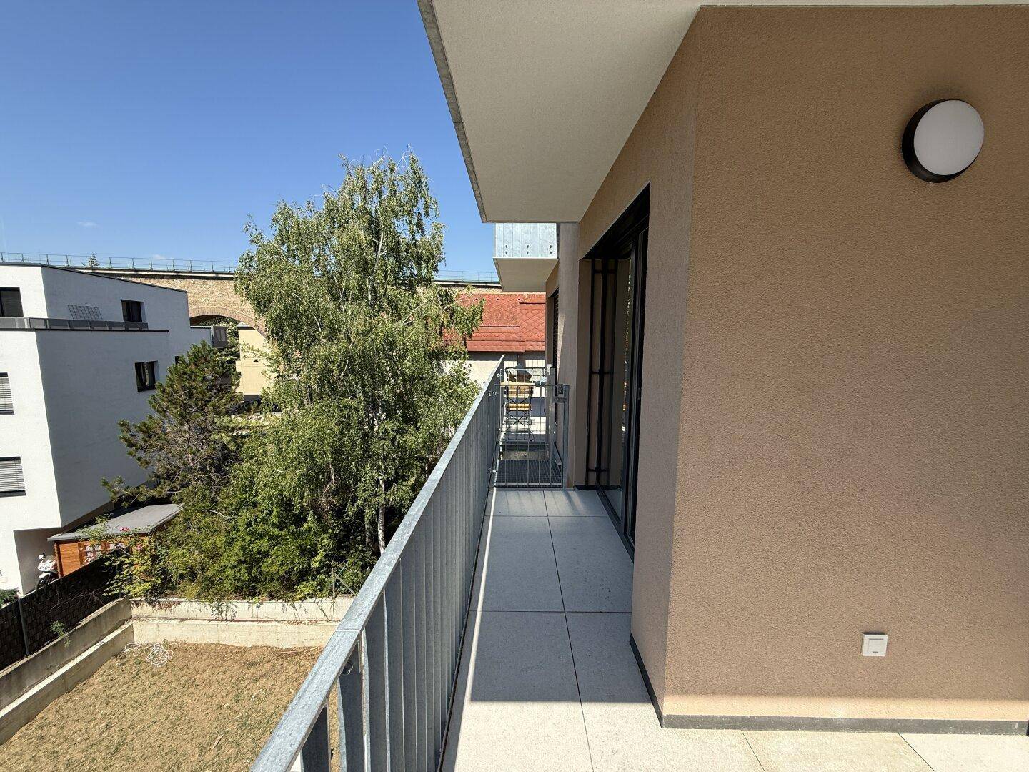 Langer Balkon mit Fliesen und Blick auf einen begrünten Innenhof und weitere Gebäude.