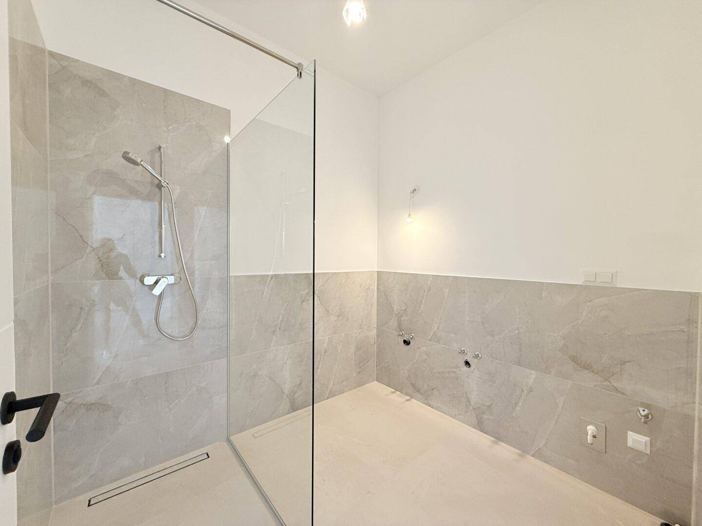 Modernes Badezimmer mit begehbarer Dusche und hellgrauen Fliesen, bereit für die Installation der Armaturen.