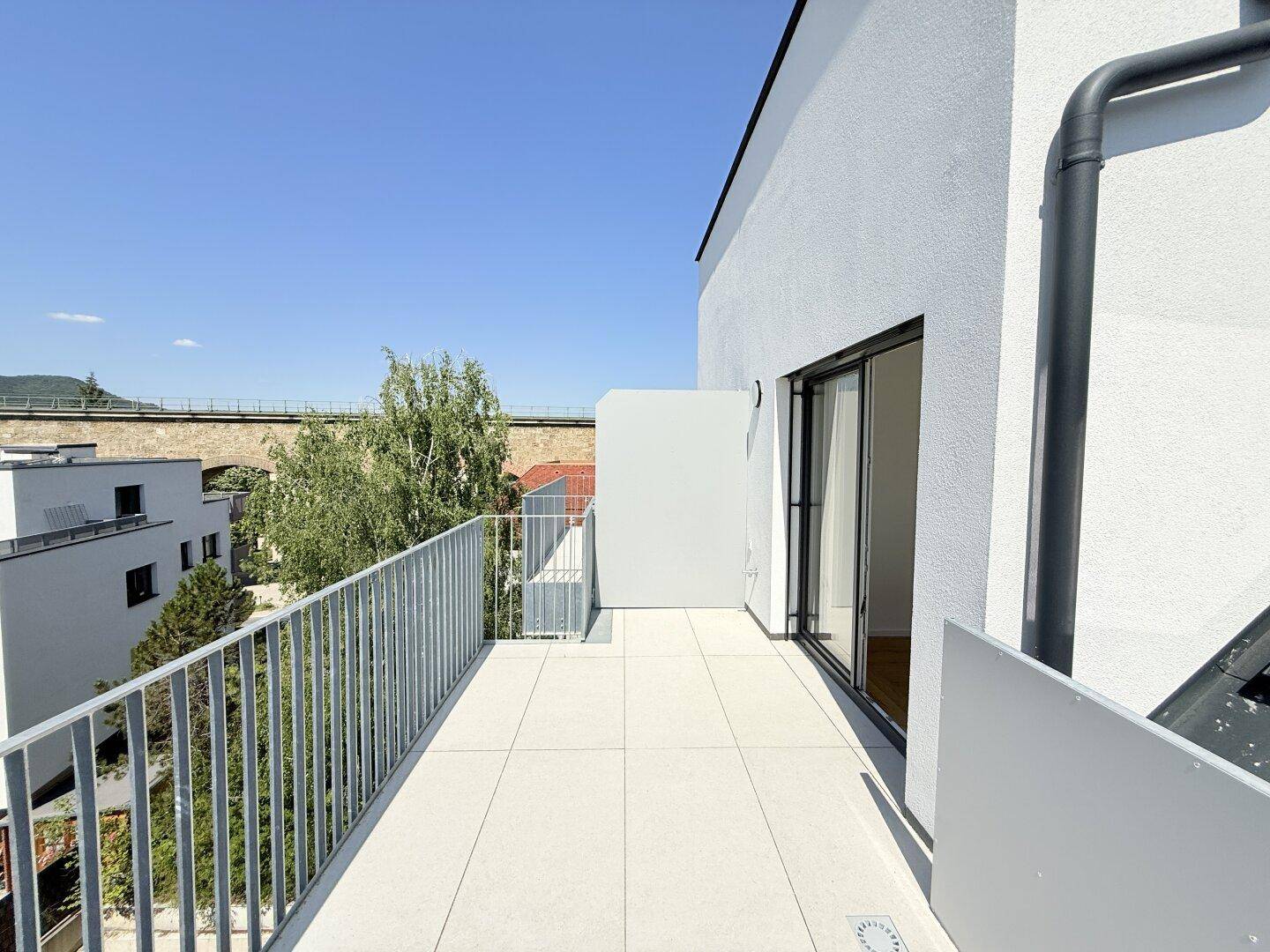 Geräumige Terrasse mit Fliesenboden und Blick auf eine Brücke sowie grüne Bäume, bietet viel Sonne.