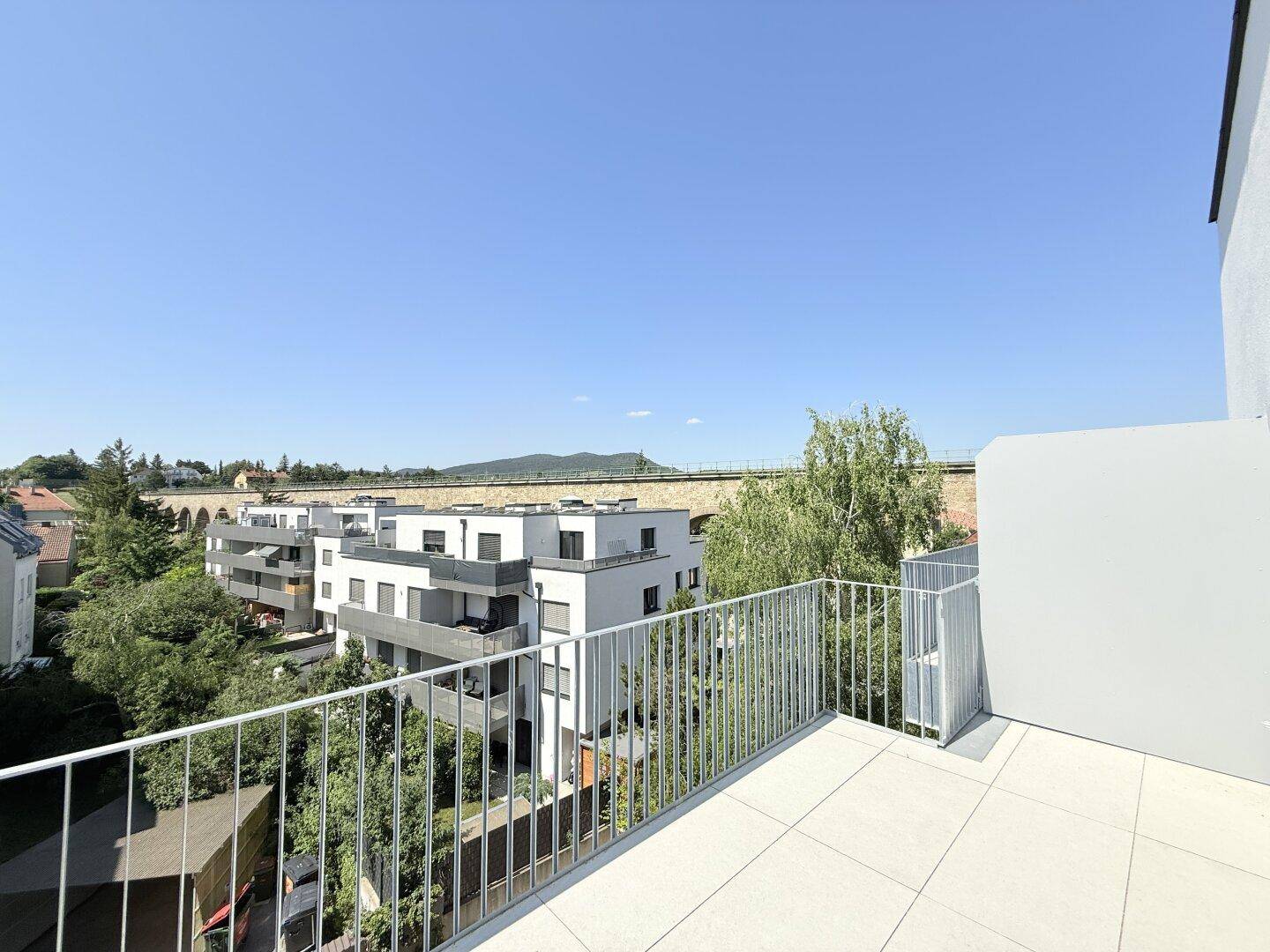 Weitläufige Terrasse mit Panoramablick über moderne Gebäude und eine hügelige Landschaft, sehr sonnig.