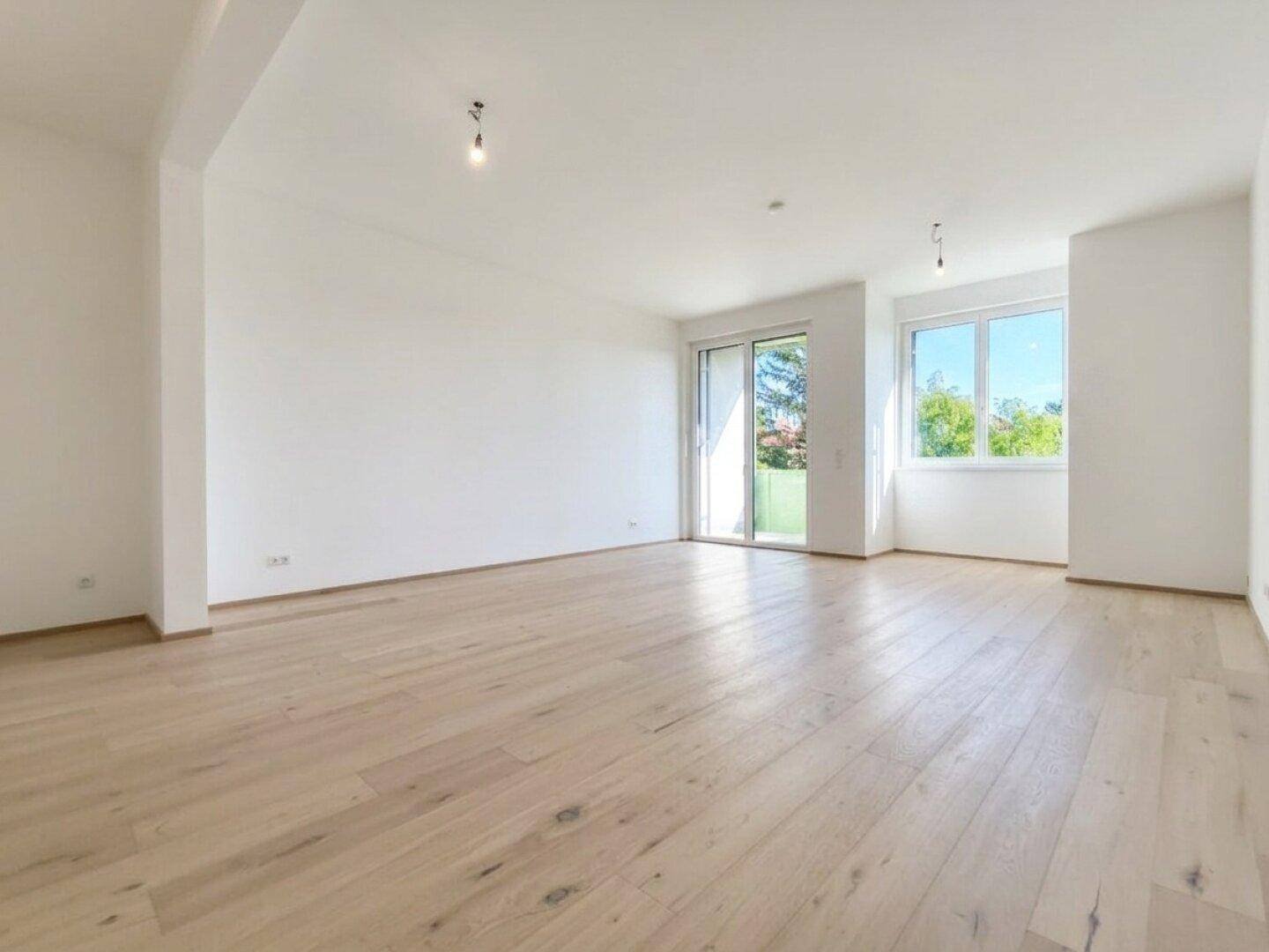 Geräumiges, leeres Wohnzimmer mit hellem Holzboden und großen Fenstern.