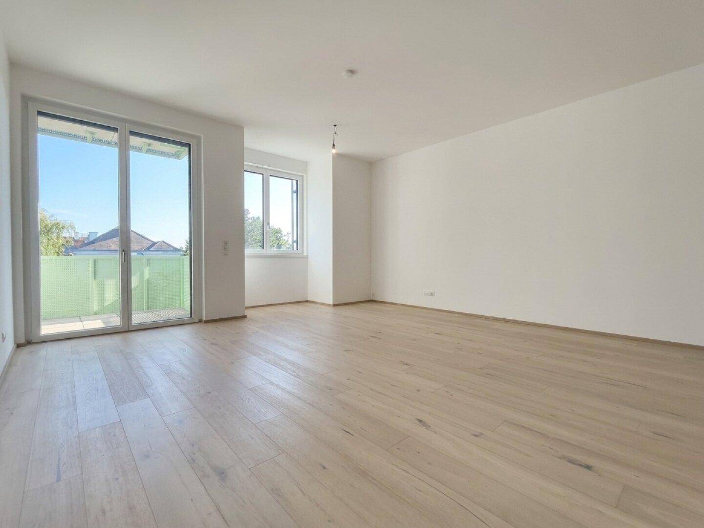 Unmöbliertes Wohnzimmer mit Holzboden, Balkonzugang und Blick ins Grüne.
