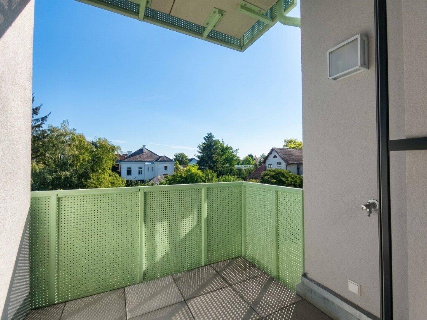 Heller Balkon mit grüner Brüstung und Blick auf die umliegenden Häuser und Bäume.