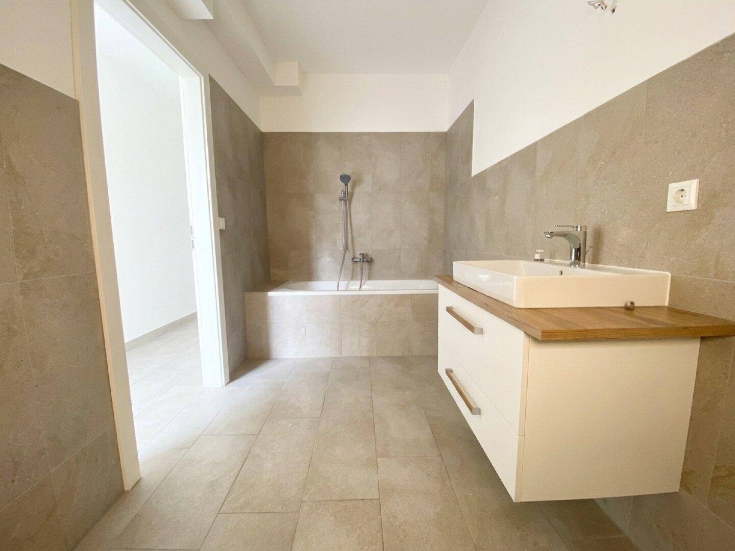Modernes Badezimmer mit Badewanne, schwebendem Waschtisch und beigefarbenen Fliesen in neuwertigem Zustand.