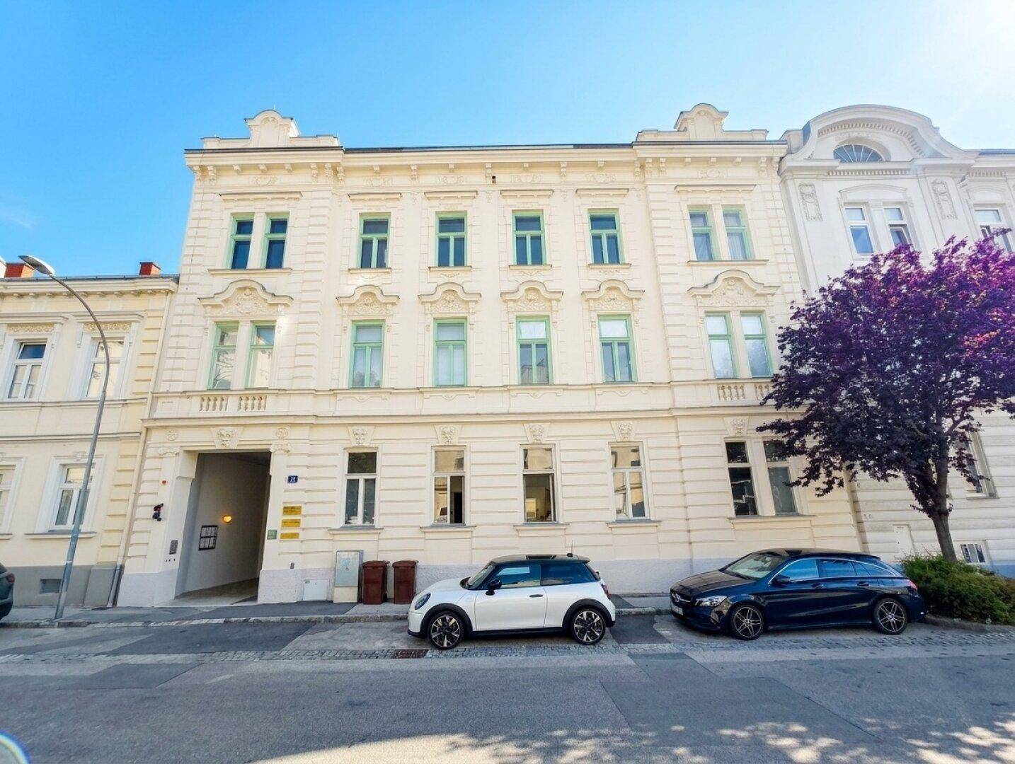 Historische Fassade eines Altbaus an einer Straße mit parkenden Autos und Bäumen.