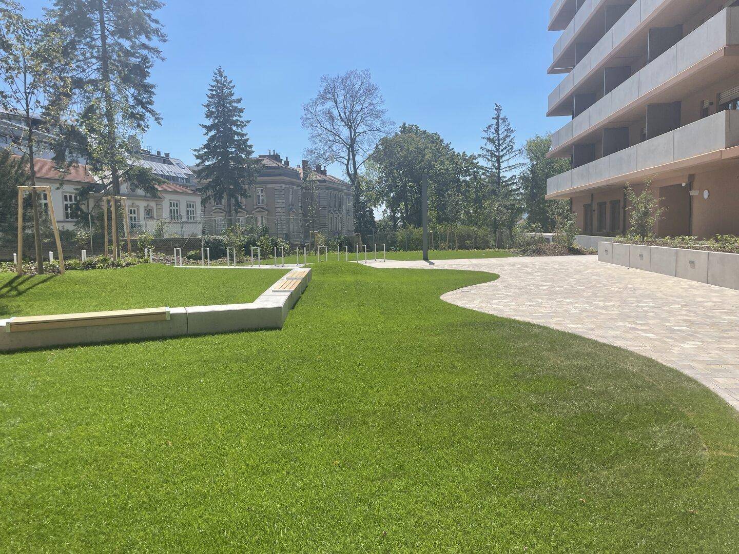 Gepflegte Grünfläche mit Rasen, Sitzbänken und Fahrradständern, umgeben von moderner Architektur.