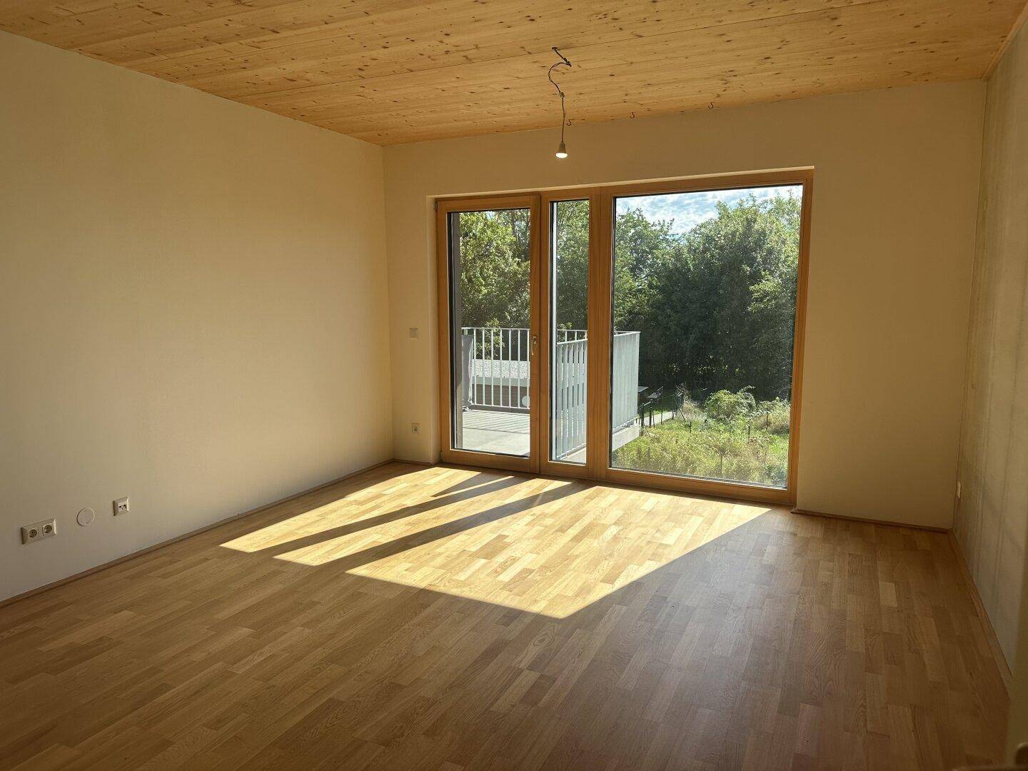 Heller Raum mit Holzboden und Holzdecke, großen Fenstern und Zugang zum Balkon mit Grünblick.