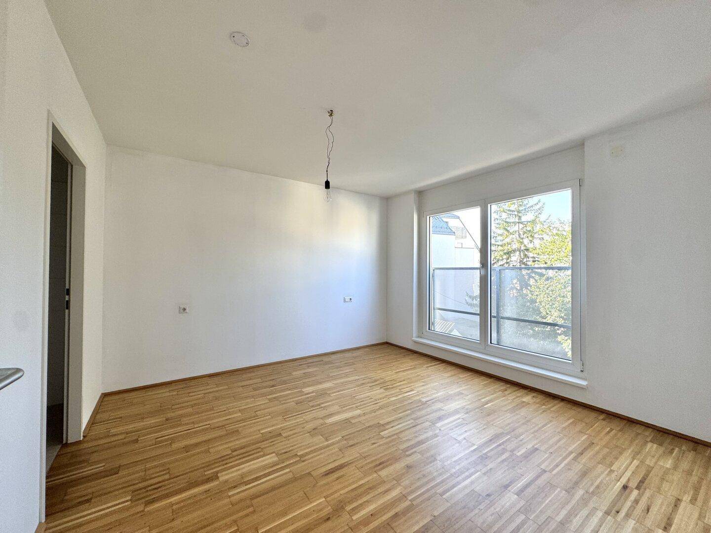 Geräumiges Zimmer mit hellem Parkettboden und einem großen Fenster, das den Raum erhellt.