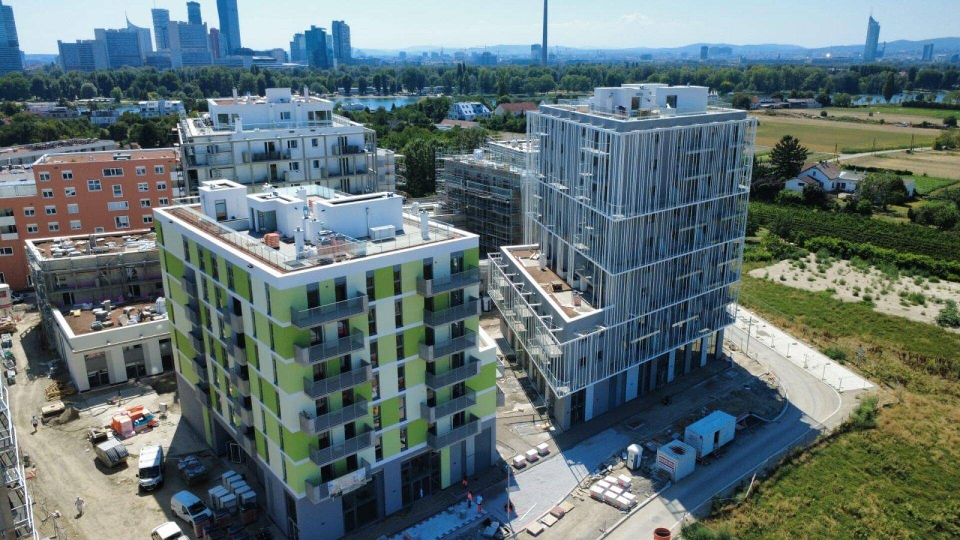 Luftaufnahme von Neubauten mit grünen und grauen Fassaden, im Hintergrund die Wiener Skyline.