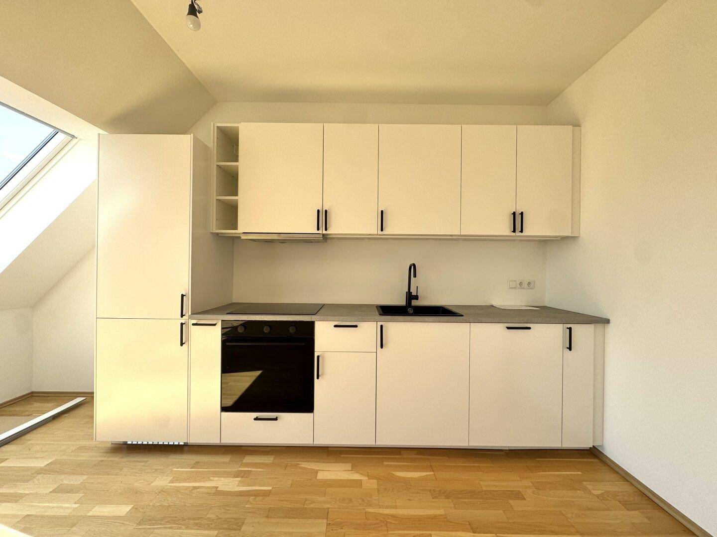 Moderne Küchenzeile mit weißen Schränken, integriertem Backofen und dunkler Arbeitsplatte in hellem Raum.