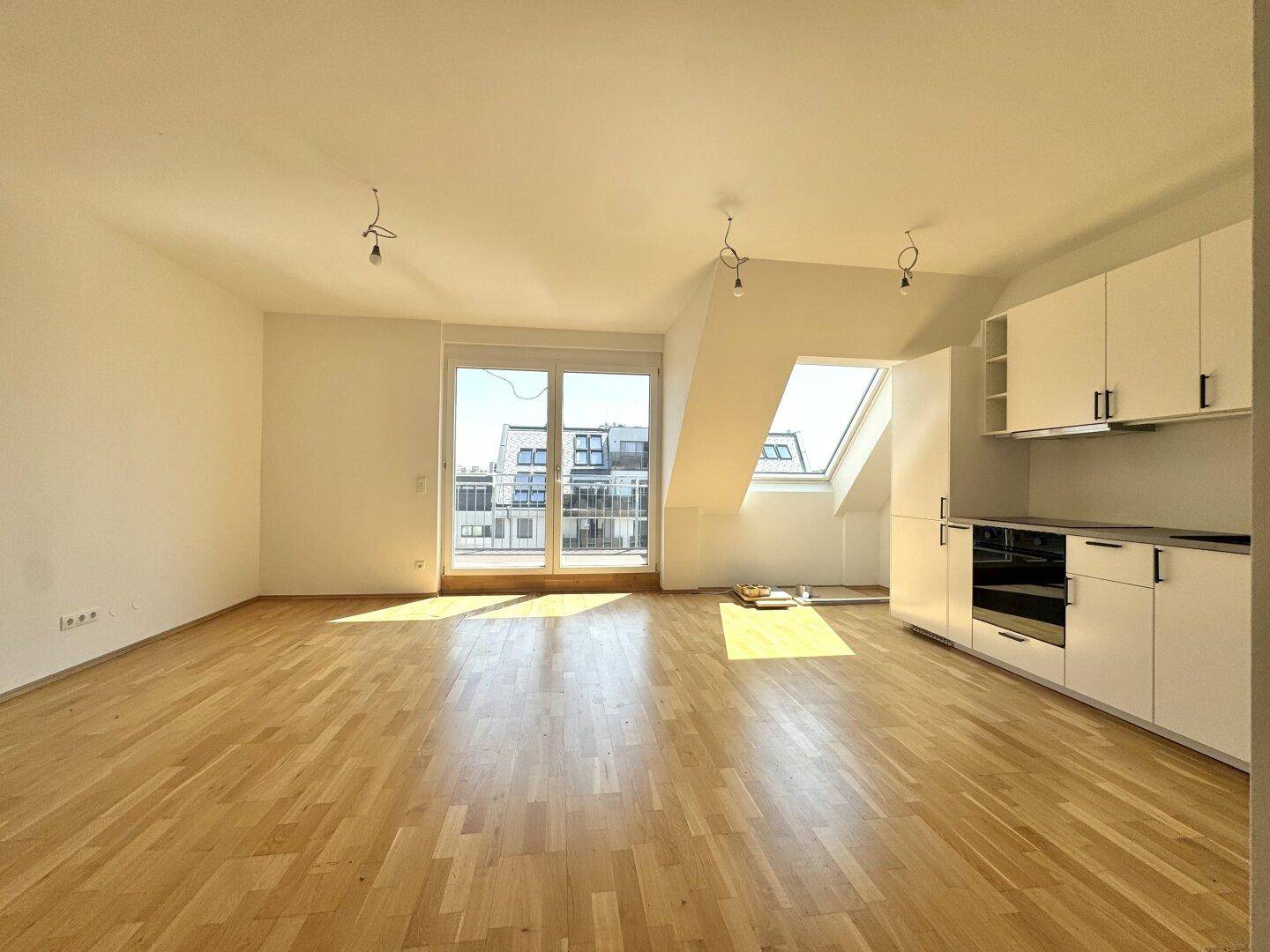 Heller Wohnbereich mit Holzboden, Dachschrägen und integrierter Küchenzeile, bietet Zugang zum Balkon.