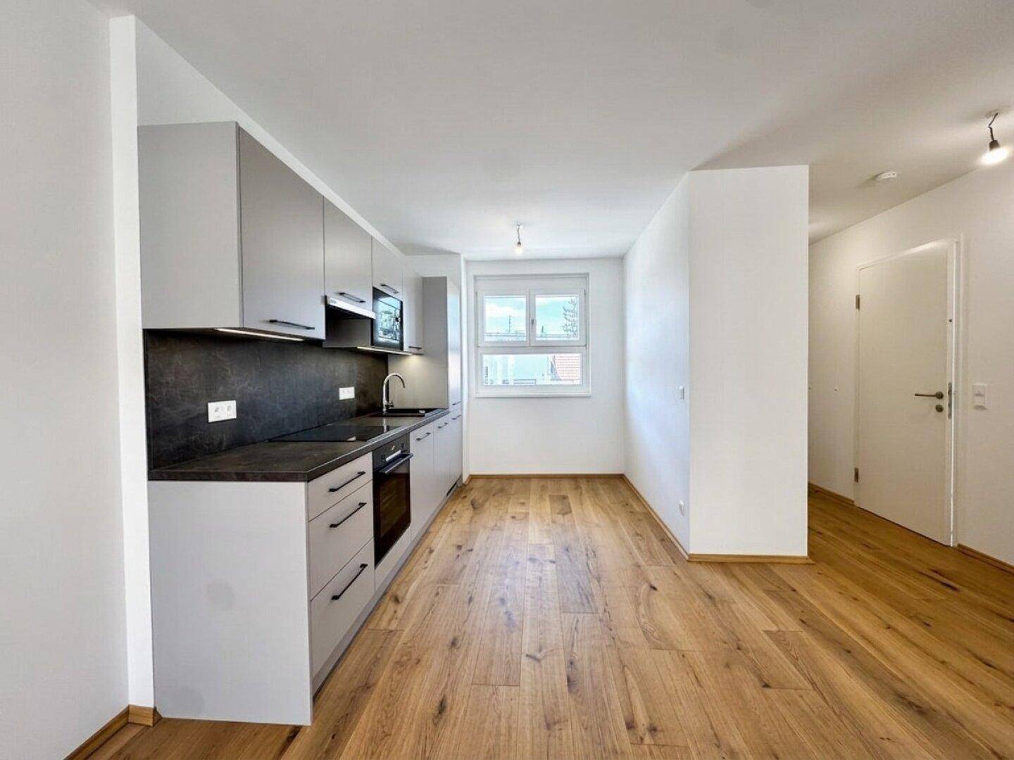 Moderne Küche mit hellgrauen Schränken, dunkler Arbeitsplatte und Holzboden, mit Fenster.