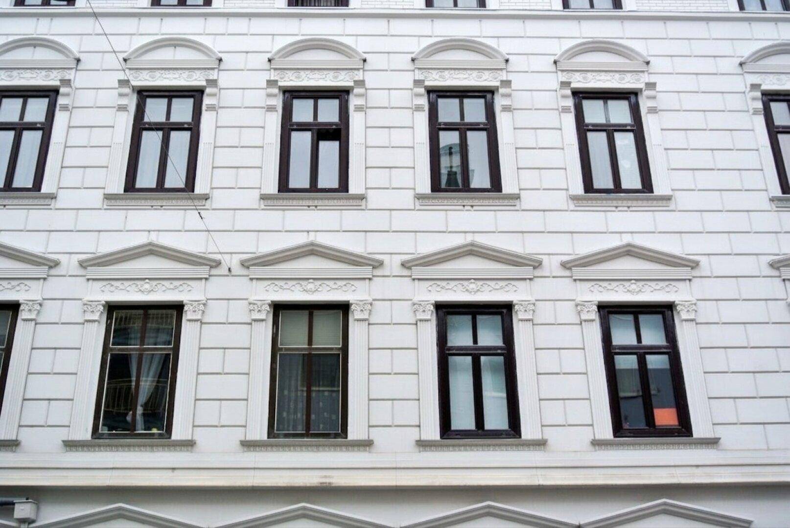 Detailansicht der weißen Fassade eines Altbaus mit verzierten Fenstern und braunen Rahmen.