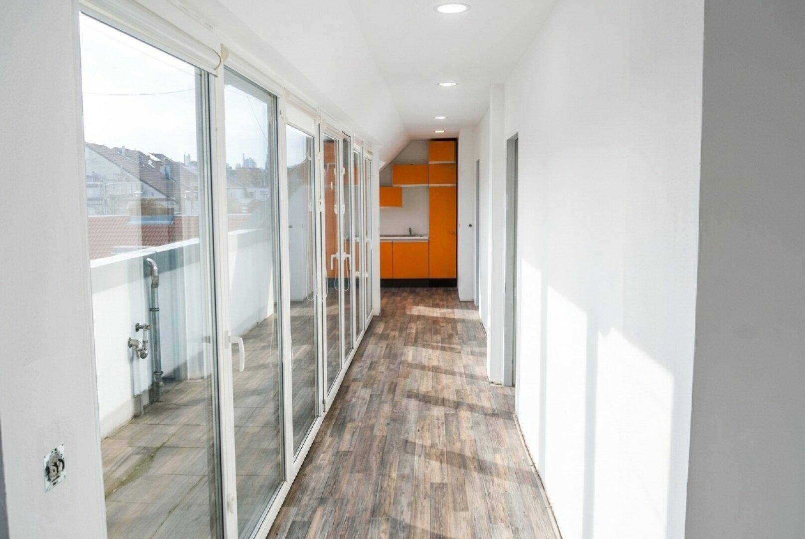 Heller Flur mit Holzboden und Zugang zu einem Balkon, im Hintergrund eine orangefarbene Küche.