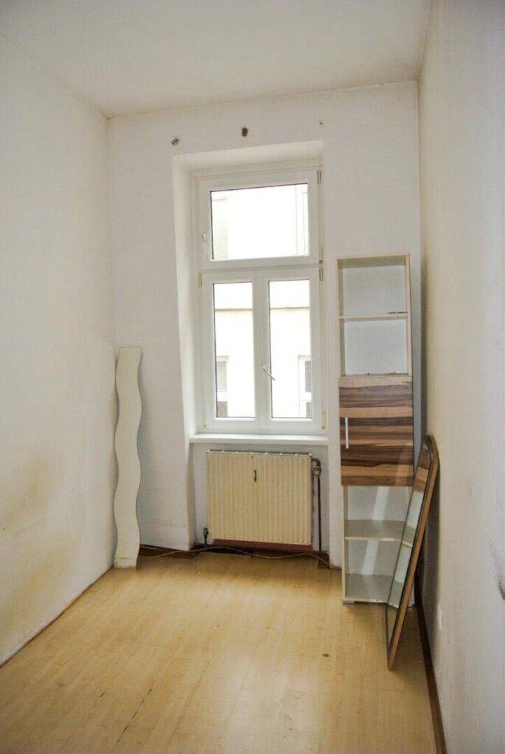 Leeres Zimmer mit weißen Wänden, hellem Holzfußboden und einem Fenster mit Heizkörper.