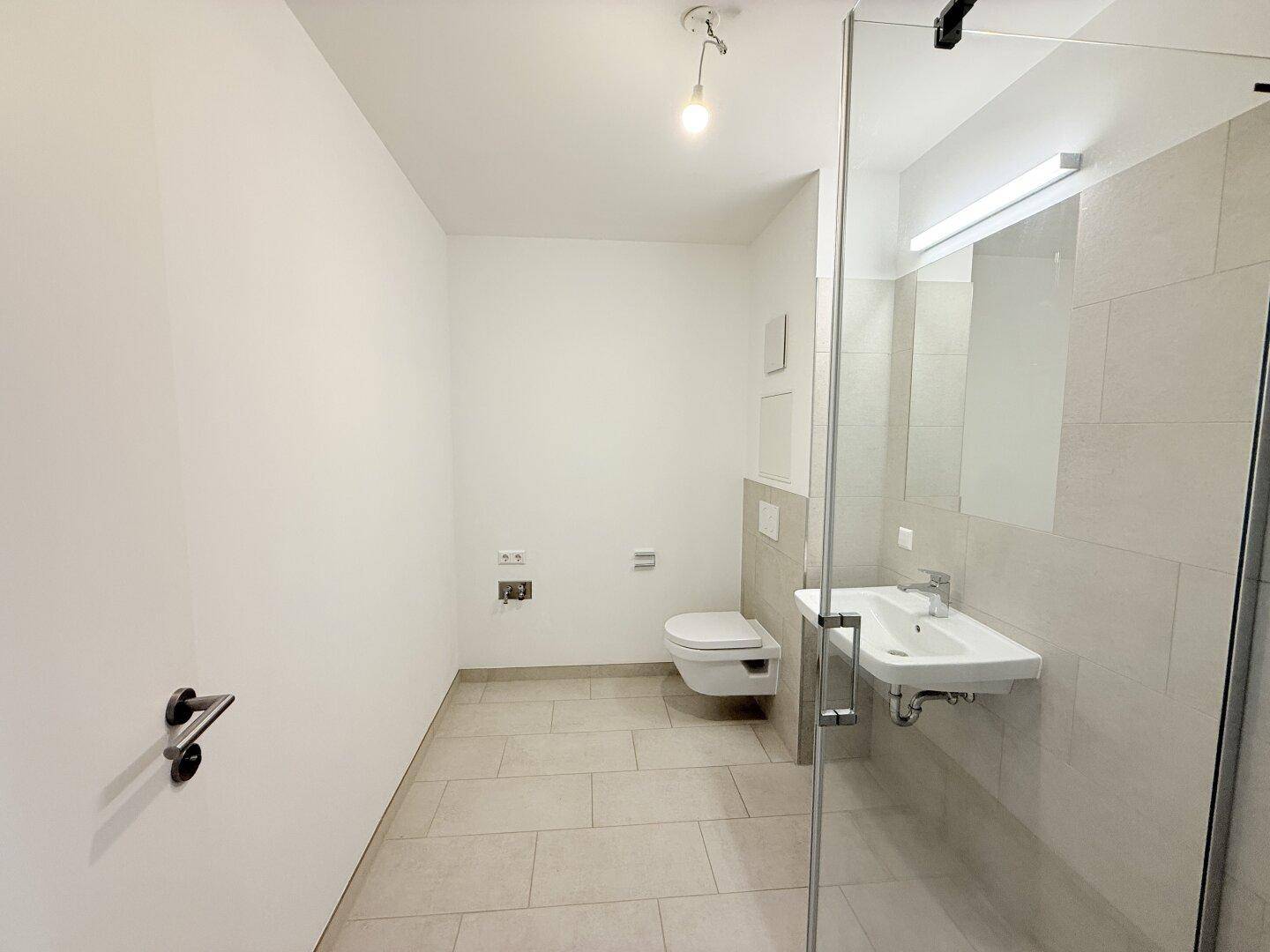 Modernes Badezimmer mit Duschkabine, wandhängendem WC und Waschbecken, in hellen Fliesen gehalten.