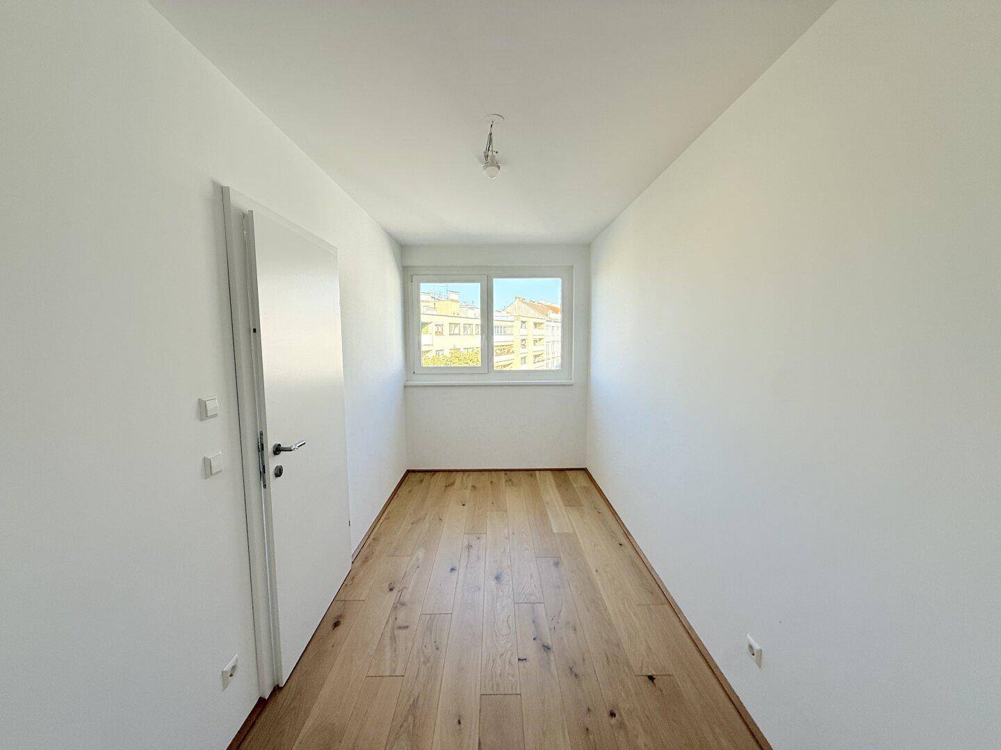 Helles Zimmer mit Fenster, Holzboden und weißen Wänden, bietet vielseitige Nutzungsmöglichkeiten.