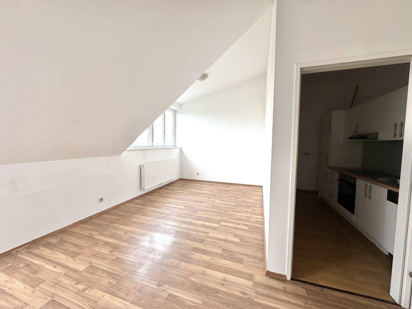 Heller Wohnbereich mit Laminatboden und Dachschräge, angrenzend an eine kleine Küche.