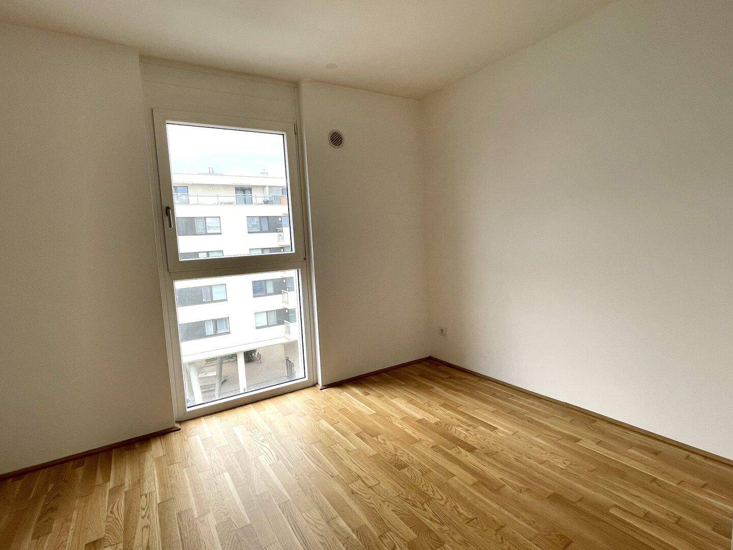 Helles Schlafzimmer mit Parkettboden und großem Fenster, das viel Tageslicht hereinlässt.