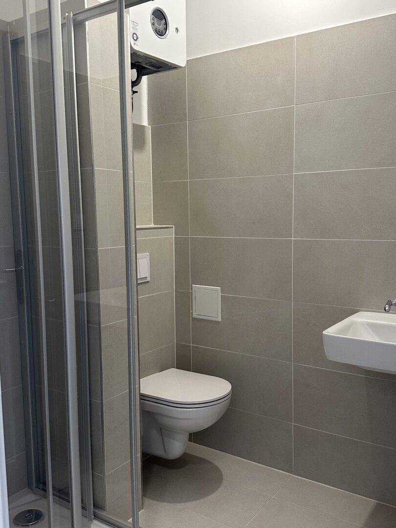 Modernes Badezimmer mit grauen Fliesen, Duschkabine und wandhängendem WC.