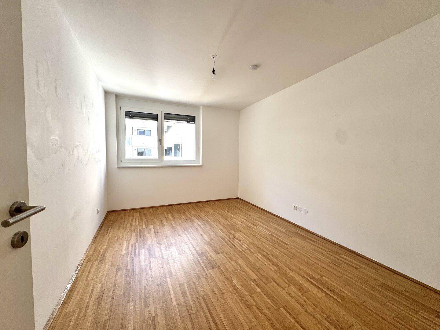 Helles Schlafzimmer mit Parkettboden und einem Fenster, das viel Tageslicht hereinlässt.