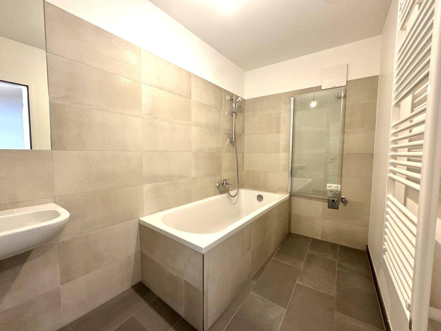 Modernes Badezimmer mit beigefarbenen Wandfliesen, Badewanne mit Duschfunktion und Handtuchheizkörper.
