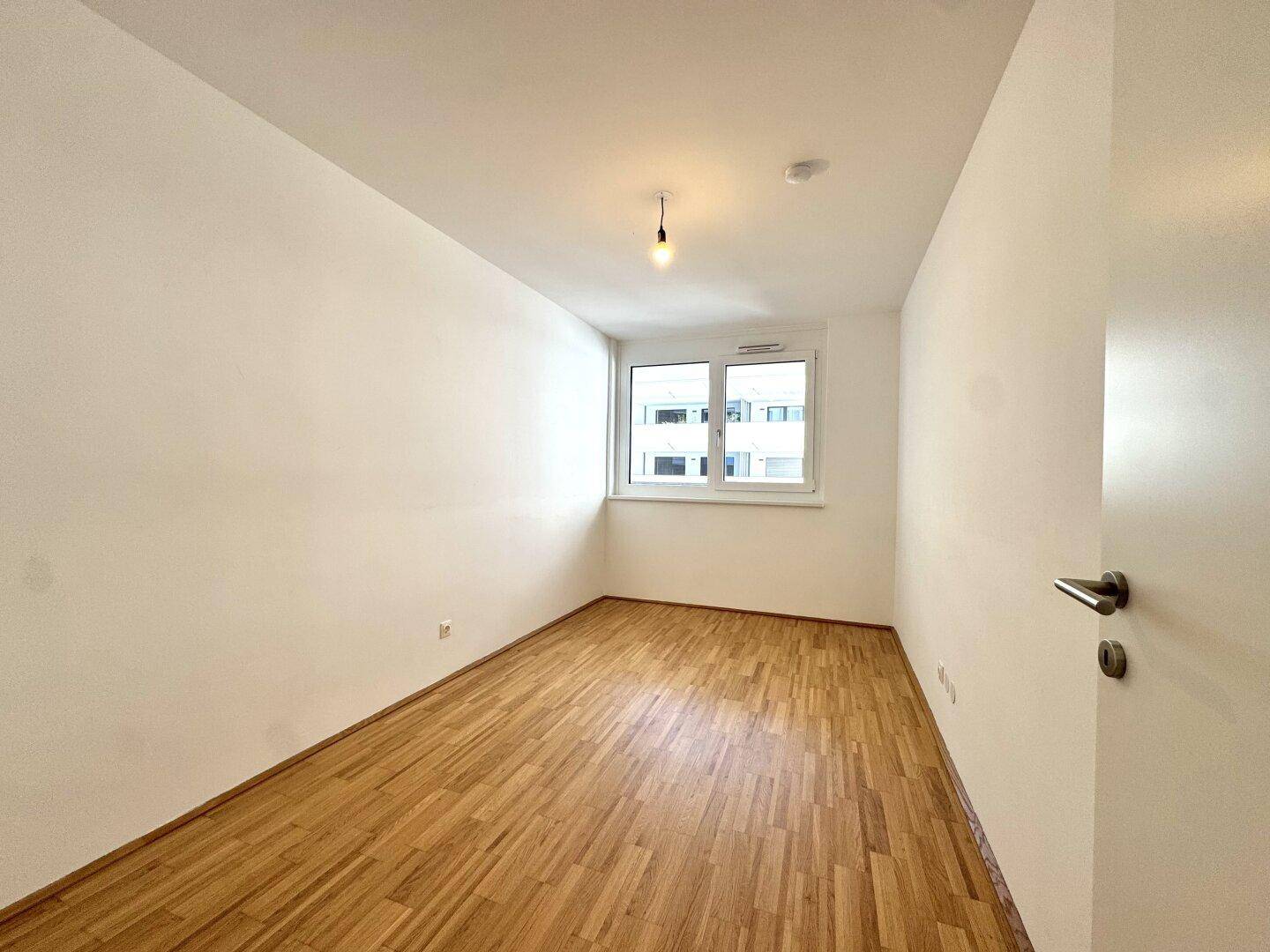 Freundliches Schlafzimmer mit hellem Parkettboden und einem Fenster mit Blick auf die Umgebung.
