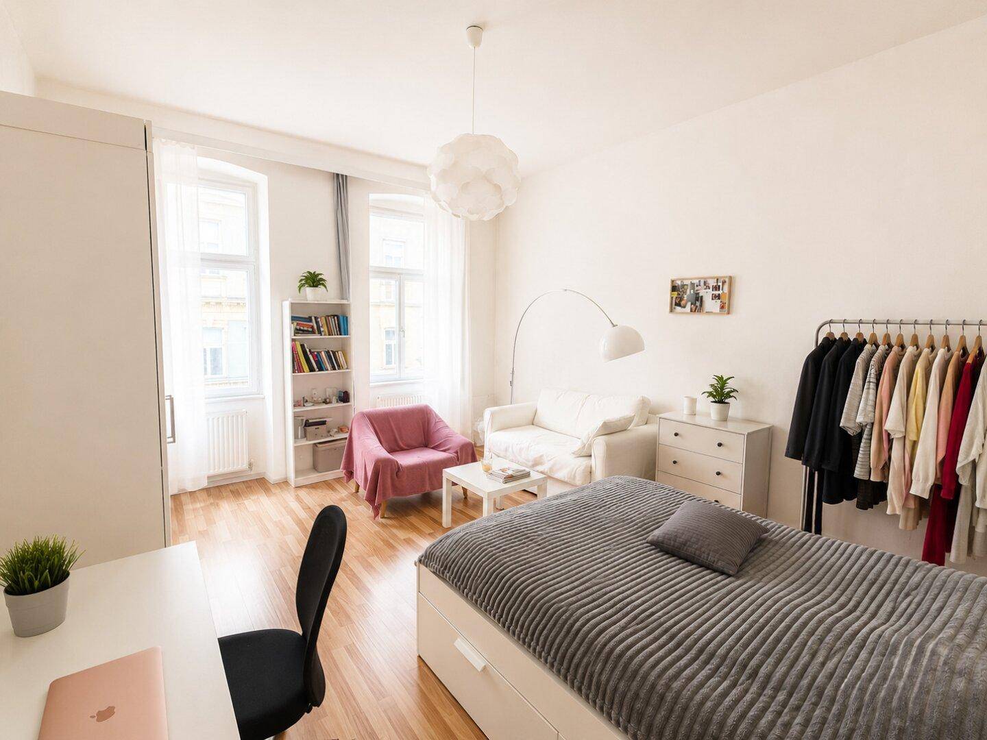 Freundliches Schlafzimmer mit Holzboden, Bett, Sofa und Kleiderstange, gut beleuchtet.