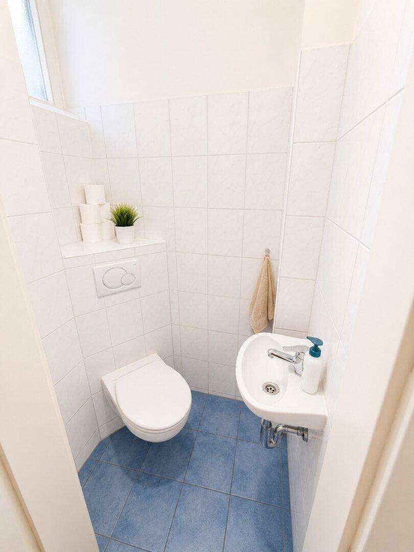 Separates WC mit kleinem Waschbecken, weißen Wandfliesen und blauen Bodenfliesen.