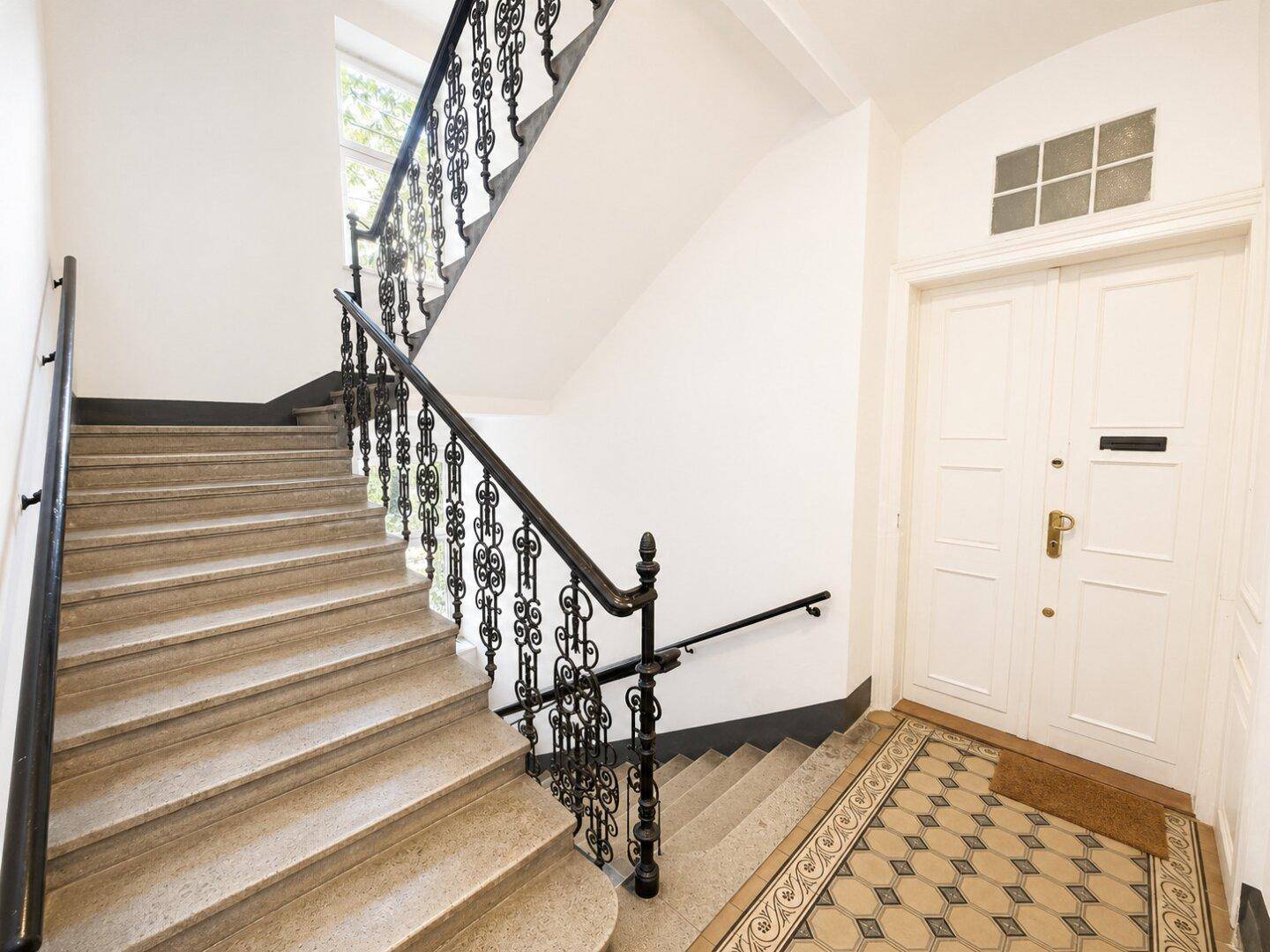 Historisches Treppenhaus mit Steinstufen, schmiedeeisernem Geländer und gemustertem Boden.