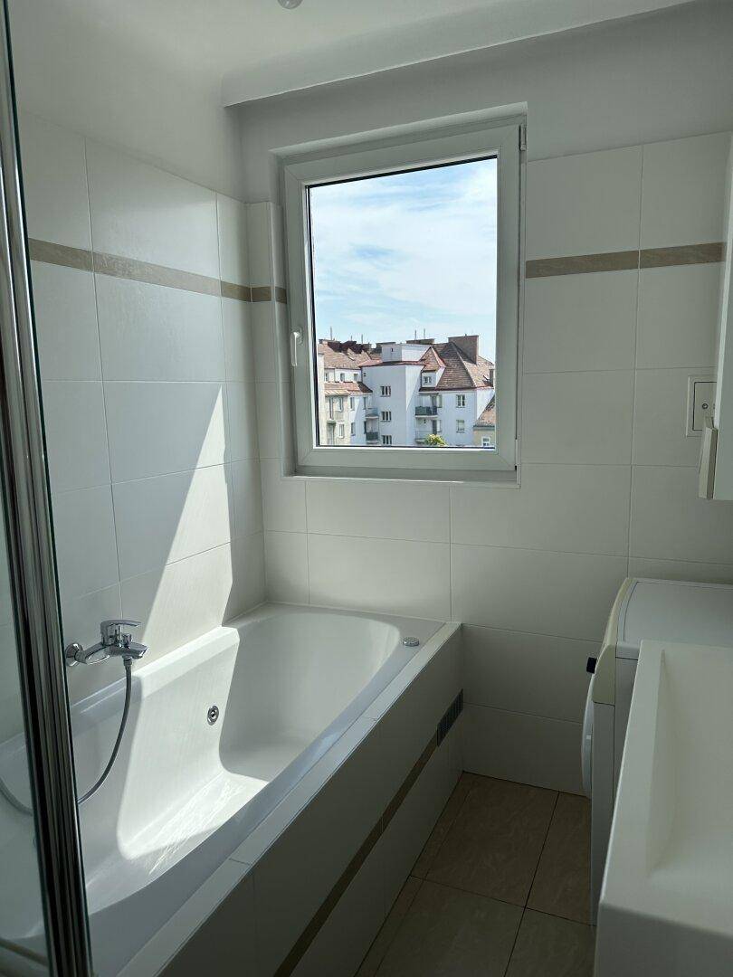 Helles Badezimmer mit Badewanne, weißen Fliesen und einem Fenster mit Stadtblick.