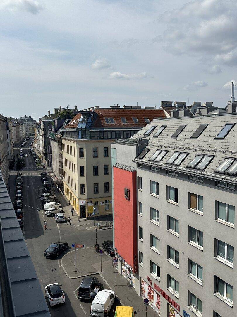 Blick von einem Balkon auf eine belebte Stadtstraße mit Gebäuden und parkenden Autos.