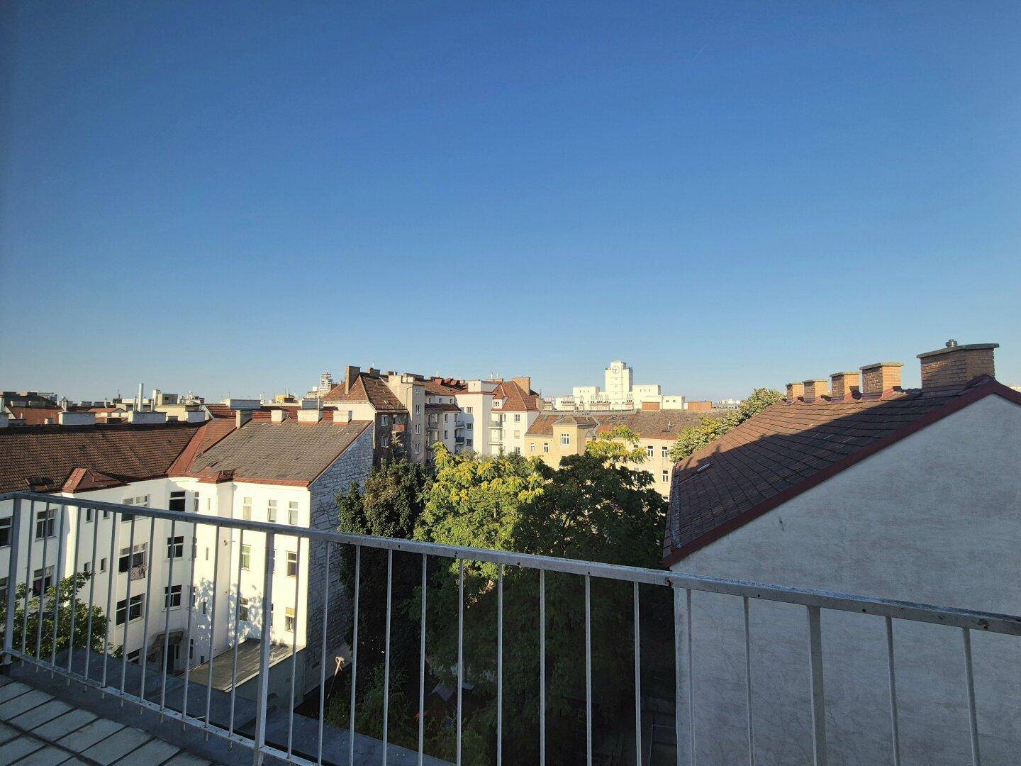 Weitläufiger Blick über die Dächer der Stadt mit grünen Bäumen unter klarem blauem Himmel.