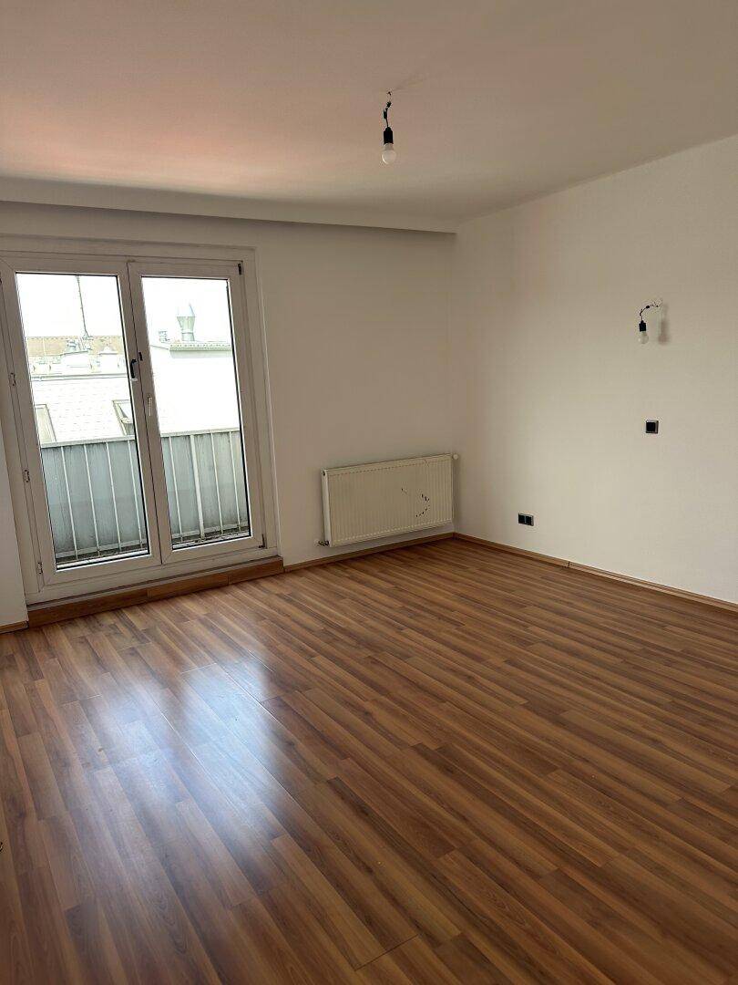 Leeres Zimmer mit Laminatboden, Heizkörper und Zugang zu einem kleinen Balkon.