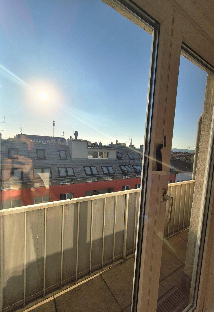 Sonniger Blick vom Innenraum auf einen Balkon mit weißem Geländer und Stadtdächern.