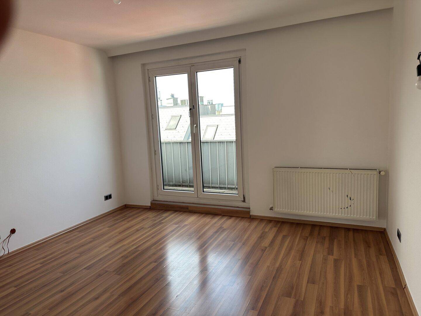 Leeres Zimmer mit Laminatboden, Heizkörper und Zugang zu einem kleinen Balkon.