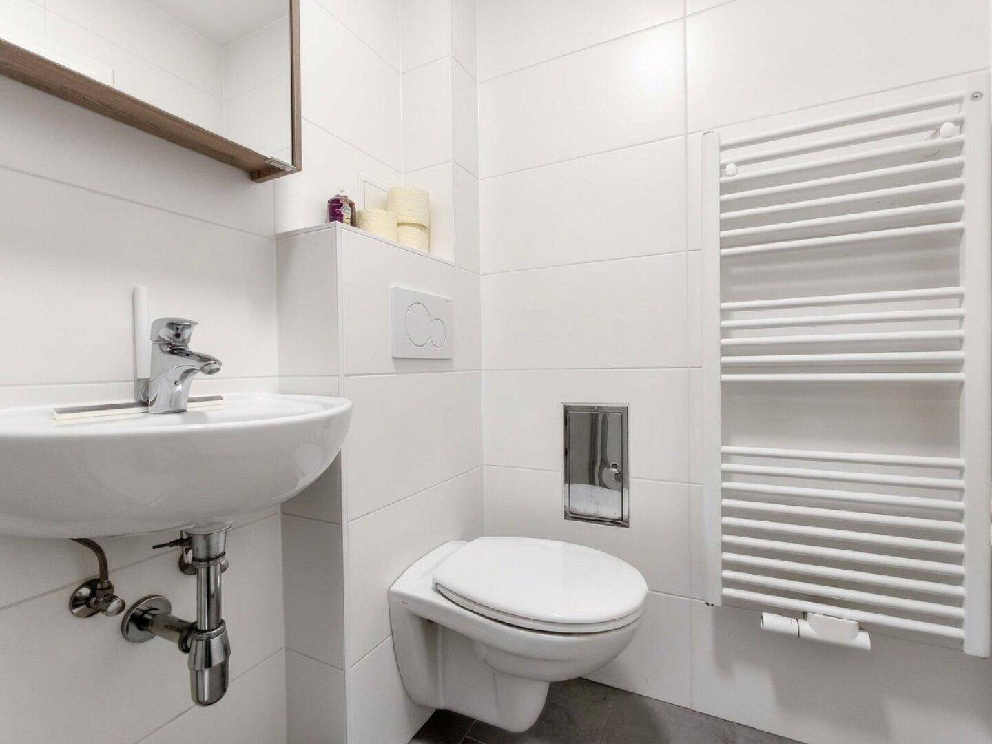 Badezimmer mit wandhängendem WC, Waschbecken, beheiztem Handtuchhalter und weißen Fliesen.