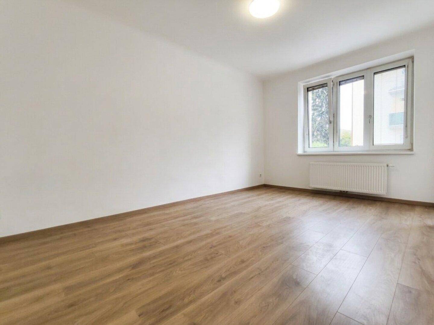 Leeres Schlafzimmer mit hellem Laminatboden und großem Fenster, das viel Tageslicht hereinlässt.