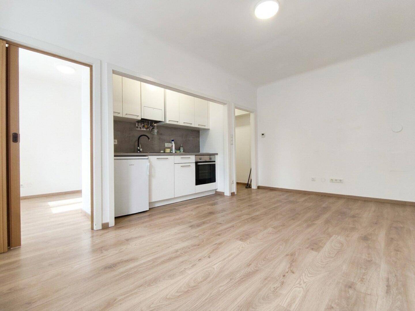 Offener Wohnbereich mit Küchenzeile und Durchgang zu einem angrenzenden Zimmer, mit Laminatboden.